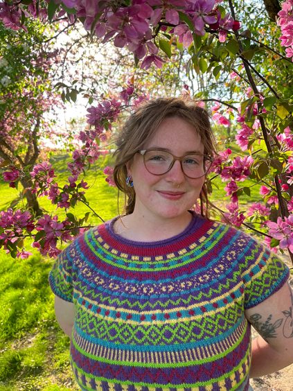 Margo Roberts in a colorful knit sweater, standing under a flowering tree