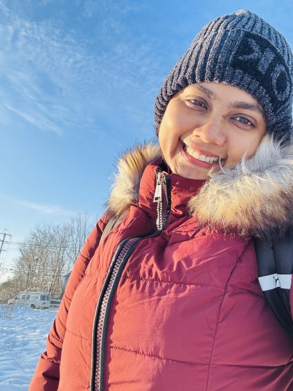 Nahida Kabir in winter coat and hat, smiling at the camera, with snow in the background and a blue sky