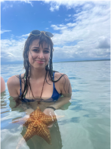 Young woman in swimsuit and sunglasses standing chest-deep in the ocean, holding a starfish in front of her