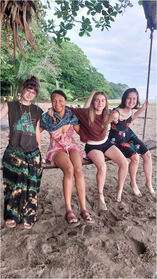 Four young women sitting side by side on a long swinging bench near a beach