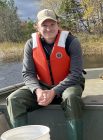 Shawn Snyder sitting in a boat with a lifejacket on, hands folded in lap, grinning. The boat is in a pond.