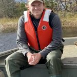 Shawn Snyder sitting in a boat with a lifejacket on, hands folded in lap, grinning. The boat is in a pond.