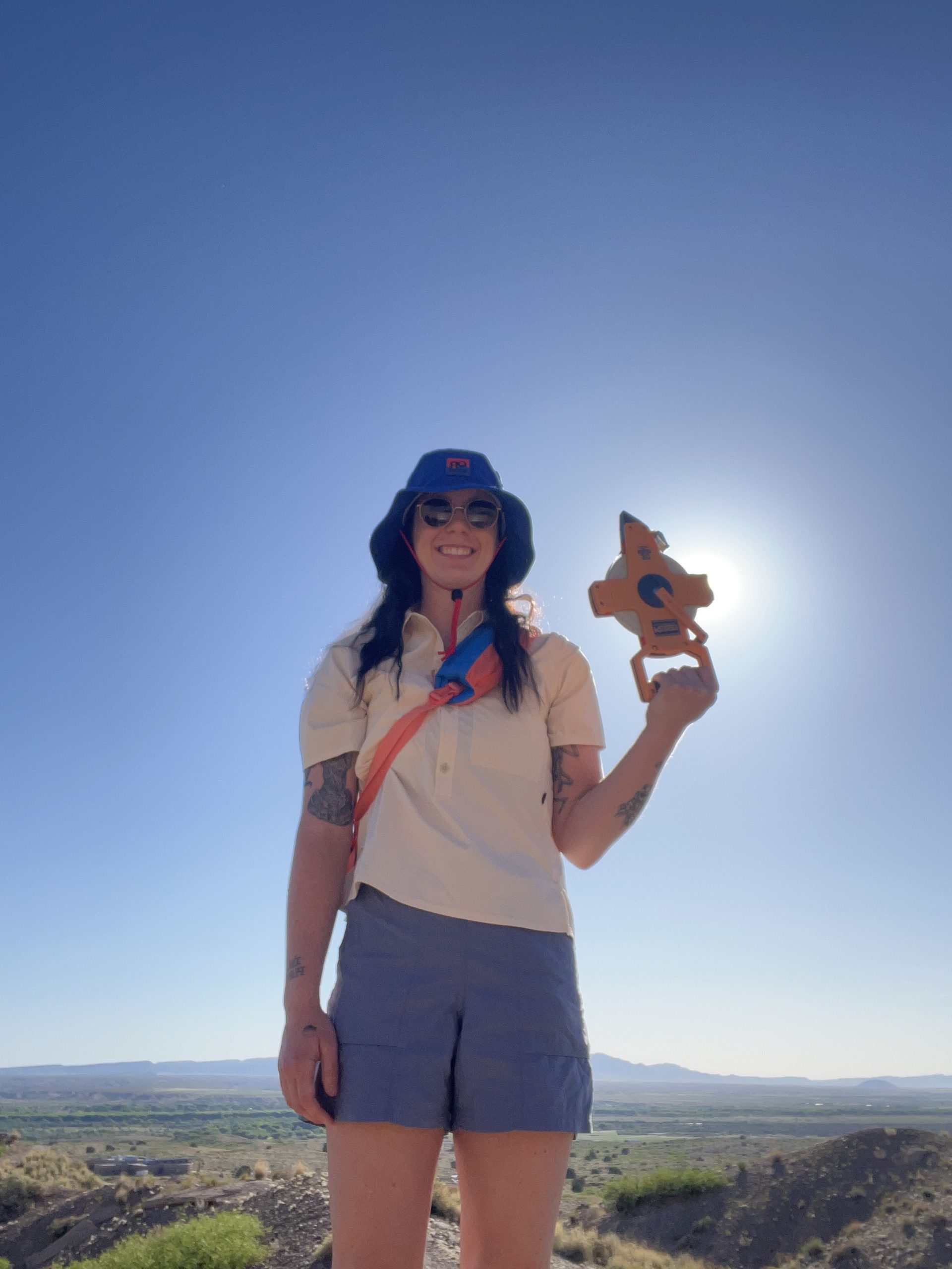 Looking up at Emily Kanach, who is standing in front of the sun, smiling, wearing a floppy hat and sunglasses