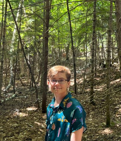 Salem Ermish wearing a shirt with insects printed on it, standing in the woods, with a deer in the distance