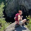 Mary Rose Holland squatting on top of a rocky outcrop, wearing a backpack, and giving a thumbs-up