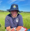 Kayla McLagan wearing a floppy hat, looking down and smiling, sitting in a field of grasses under a blue sky