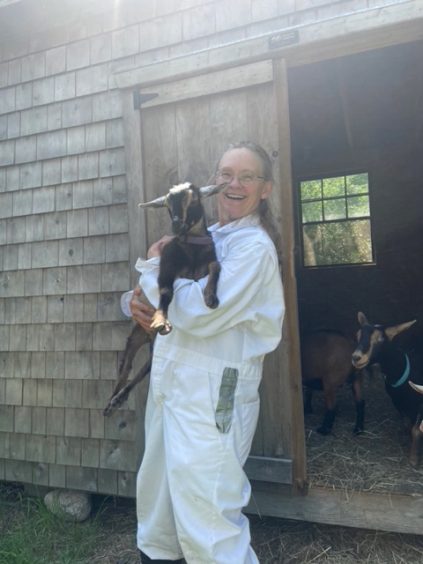 Woman with long gray hair in a ponytail, wearing glasses and white work jumpsuit, holding a goat by a barn